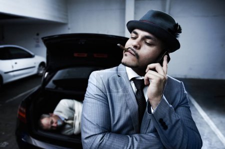 Photo for Phone call, car and criminal with hostage in trunk for negotiation, kidnapping danger and crime. Mafia, gangster and business man smoking in boot for abduction, crisis and ransom in parking lot. - Royalty Free Image