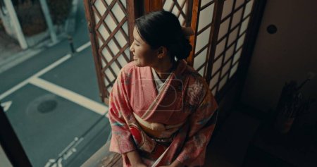 Foto de Mujer japonesa, vista a la ventana y ropa tradicional en casa para la visión, ideas o recordar por la calle. Chica, persona o pensamiento con sonrisa, feliz memoria o atención plena en la mañana en el apartamento en Tokio. - Imagen libre de derechos