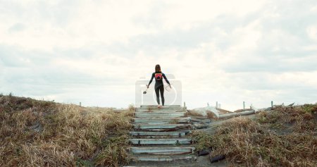 Foto de Mujer, surfista con tabla de surf en la playa y vista trasera, naturaleza y deportes extremos para la aptitud. Atleta en escaleras, ejercicio y surf, entrenamiento o entrenamiento con el océano, salud y bienestar con hobby. - Imagen libre de derechos