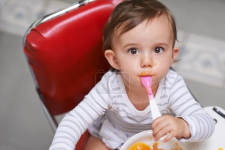 Téléchargez les photos : Manger, mignon et portrait de bébé dans la chaise avec de la nourriture végétale pour le développement de l'enfant à la maison. Enfant ou tout-petit sucré, nourrissant et affamé qui mange sainement le déjeuner, le dîner ou le souper à la maison - en image libre de droit