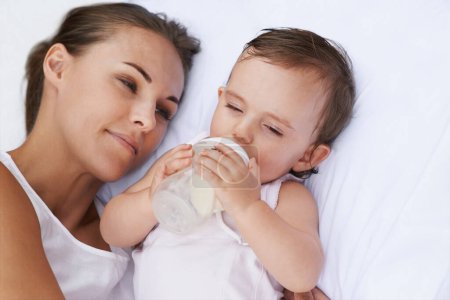 Photo for Baby, mother and drinking bottle for nutrition, liquid food and relaxing together on bed at home. Mommy, toddler and formula for health or child development in bedroom, feeding and milk for wellness. - Royalty Free Image