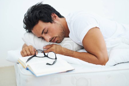 Téléchargez les photos : Dormir, lire et un jeune homme dans la chambre de sa maison avec un livre pour se détendre ou se reposer le week-end. Étudier, apprendre ou étudier avec une personne fatiguée endormie sur un livre alors qu'elle est couchée au lit. - en image libre de droit