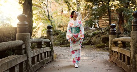 Téléchargez les photos : Marche, femme traditionnelle et japonaise dans le parc pour le calme, l'air frais et se détendre sur le pont à l'extérieur. Voyage, la culture et la personne en vêtements autochtones, le style et le kimono en vacances, la paix ou la pensée. - en image libre de droit
