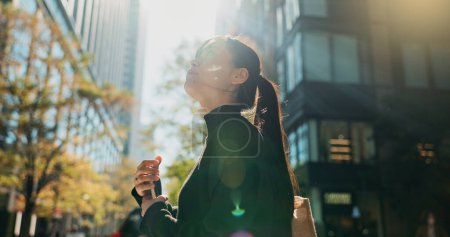 Foto de Mujer japonesa, teléfono y auriculares en la ciudad con sonrisa, caminando y escuchando música por los edificios. Persona, peatonal y smartphone con suscripción de streaming de audio con aplicación de radio en la calle del metro. - Imagen libre de derechos