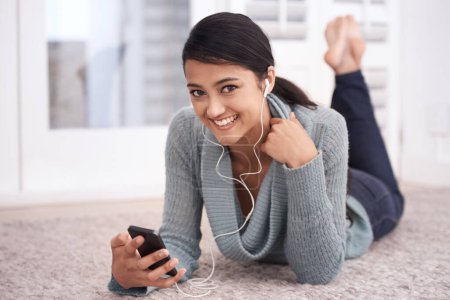 Photo for Portrait of happy woman on floor with phone, earphones and relax with mobile app for podcast in house. Face of girl lying on carpet with smile, music and streaming service fon smartphone in home - Royalty Free Image