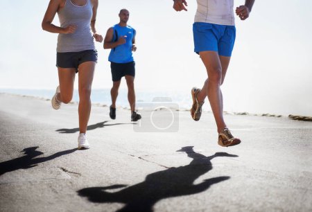 Photo for People, legs and running at beach for exercise, fitness or outdoor workout together on asphalt or road. Closeup of athletic group or runners in sports, teamwork or cardio training by the ocean coast. - Royalty Free Image