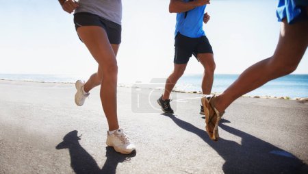 Téléchargez les photos : Les gens, la forme physique et les jambes courir à la plage pour l'exercice ou l'entraînement en plein air ensemble sur l'asphalte ou la route. Gros plan du groupe sportif ou des coureurs dans le sport, le travail d'équipe ou l'entraînement cardio au bord de l'océan. - en image libre de droit