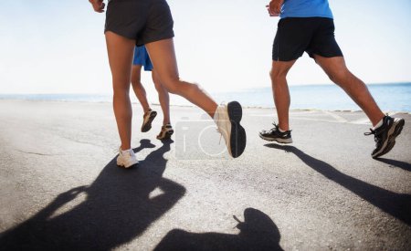 Photo for People, legs and running at beach for cardio, fitness or outdoor workout together on asphalt or road. Closeup of athletic group or runners in sports, teamwork or training exercise by the ocean coast. - Royalty Free Image