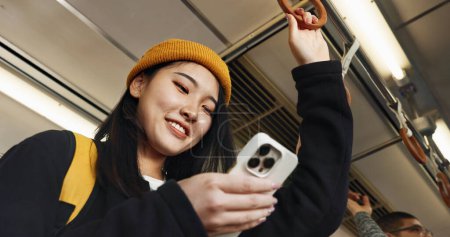 Téléchargez les photos : Femme japonaise, téléphone et SMS dans le train, sourire et voyager en cliquant, appuyez et vérifiez l'horaire sur l'application. Fille, personne et smartphone avec chat, blog de lecture et réseau social avec les transports en commun. - en image libre de droit