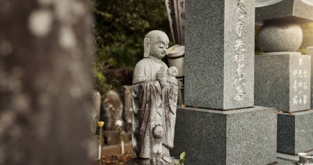 Foto de Buda, estatua y lápida en el cementerio con cultura para la seguridad, protección y escultura al aire libre en la naturaleza. Jizo, Japón y lápida conmemorativa con historia, tradición y monumento para hacer turismo. - Imagen libre de derechos