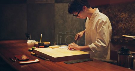 Photo for Japan chef, restaurant and cutting fish for sushi, glasses and precision with knife for healthy food in kitchen. Asian man, hands and culinary artist on job by wood board, table and raw seafood menu. - Royalty Free Image
