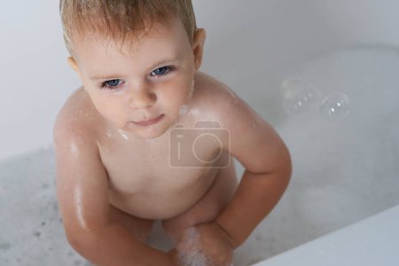 Téléchargez les photos : Bébé, enfant et bulles dans le bain d'eau pour la routine matinale, les soins de la peau et le nettoyage à la maison d'en haut. Garçon, enfant humide et tout-petit penser dans la baignoire de savon, mousse et lavage pour l'hygiène dans la salle de bain. - en image libre de droit