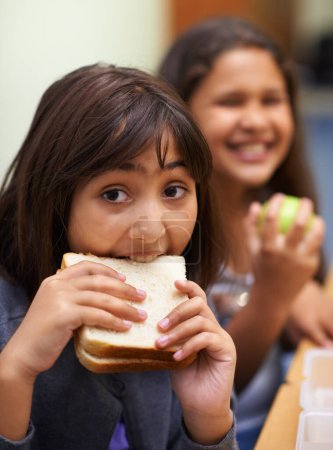 Téléchargez les photos : Fille affamée, portrait et étudiant mangeant un sandwich en classe à l'école pour les repas, pauses ou collations. Jeune enfant ou enfant élémentaire mordant du pain pour l'heure du déjeuner, des fibres ou de la nutrition en récréation de classe. - en image libre de droit