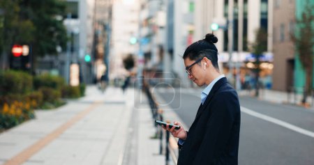 Téléchargez les photos : Téléphone, réseautage et homme d'affaires japonais dans la ville lecture email de l'entreprise sur la technologie. Carrière, voyage et professionnel jeune homme défiler ou naviguer sur un téléphone portable avec écouteurs dans la ville urbaine - en image libre de droit