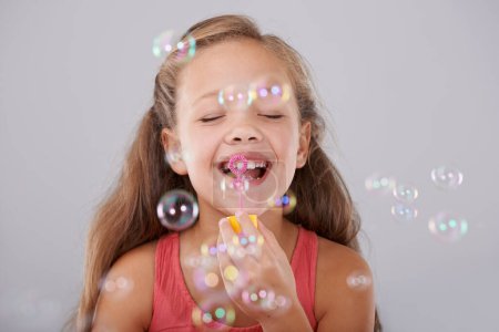 Photo for Happy, little girl and blowing bubbles in studio by toy, cute and fun games for hand eye coordination. Child, eyes closed and play with bubble wand, childhood development and smile by gray background. - Royalty Free Image