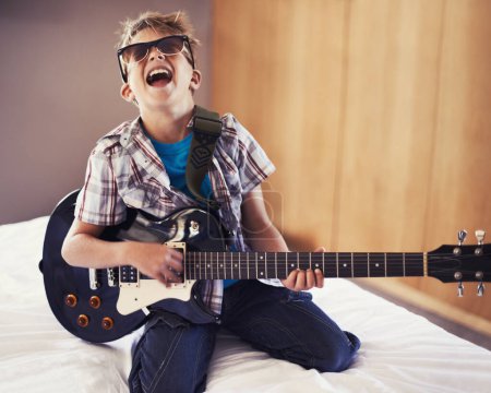Téléchargez les photos : Joyeux, jouant et enfant avec guitare sur le lit pour des leçons de musique avec des lunettes de soleil à la maison moderne. Fun, rockstar et jeune garçon enfant apprendre avec instrument à cordes électriques dans la chambre à coucher pour passe-temps à la maison - en image libre de droit