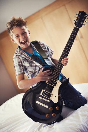 Photo for Happy, portrait and child with guitar on bed for music lesson or practice at modern home. Fun, rockstar and young boy kid playing electric string instrument for concert in bedroom for hobby at house - Royalty Free Image