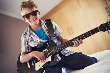 Photo for Smile, playing and child with guitar on bed for music lesson with sunglasses at modern home. Fun, rockstar and young boy kid learning with electric string instrument in bedroom for hobby at house - Royalty Free Image