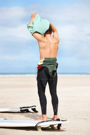 Photo for Surfing, man and getting ready with surfboard on beach with wetsuit, blue sky and dressing with mock up space. Rear view, person and athlete by ocean for training, surfer workout and extreme sports. - Royalty Free Image