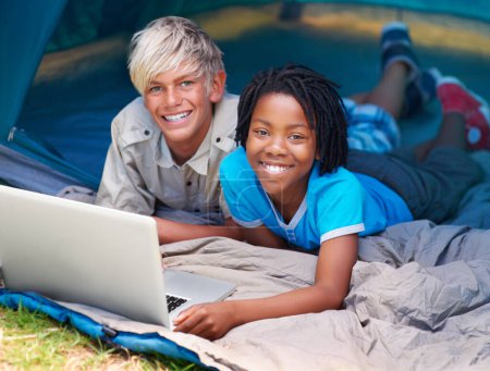 Téléchargez les photos : Enfants, garçons et heureux avec ordinateur portable pour tente de camping, médias sociaux et film en ligne avec portrait dans la nature. Amis, visage et enfants avec sourire en plein air sur l'herbe pour le jeu, se détendre et plaisir de vacances. - en image libre de droit