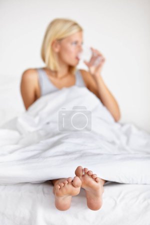 Photo for Young woman, drinking water and feet on bed for hydration, energy and health body in blanket for wellness. Blonde person, relax or natural cold drink for balance to detox, morning or liquid in studio. - Royalty Free Image