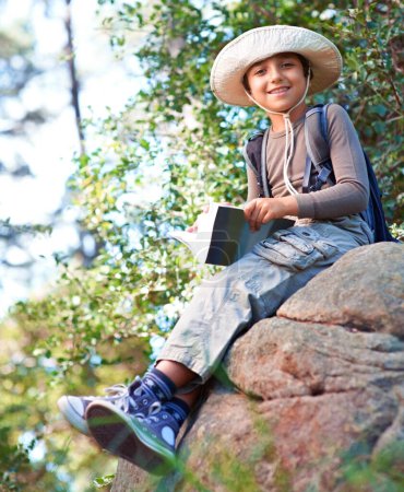 Foto de Niño, niño y senderismo por roca y libro para leer, mochilero y aventura al aire libre. Estudiante en excursión escolar, relajarse en la naturaleza y el bosque para el descubrimiento o la exploración, sonrisa y retrato de viaje. - Imagen libre de derechos