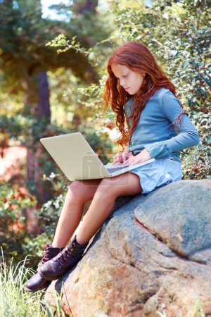 Photo for Young girl, forest and laptop on rock for internet, break and connection on holiday in woods for travel tips. Child, red hair and computer on boulder for online research, camping and relax by trees. - Royalty Free Image