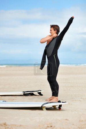 Foto de Surfista, hombre y preparándose con tabla de surf por el océano con traje de neopreno, cielo azul y vestirse con simulacro de espacio. Deportes extremos, deportista y persona en la playa para entrenar, practicar surf y aventura. - Imagen libre de derechos