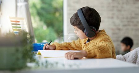 Photo for Boy child, homework and writing with headphones, music and focus for learning, drawing or education. Young male kid, notebook and study with audio streaming, radio or listening online in family house. - Royalty Free Image