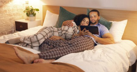 Photo for Couple, happy and relax in bedroom with tablet at night, streaming movie and hug. Smile, technology and man and woman in bed on social media app, watching online video and bonding together in home - Royalty Free Image