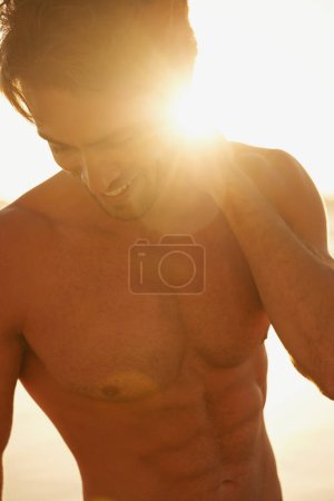 Photo for Man, shirtless and happy on beach at sunset, confidence and tourist at sea on summer holiday. Male person, lens flare and outdoor adventure for fun, calming ocean and peaceful environment in nature. - Royalty Free Image