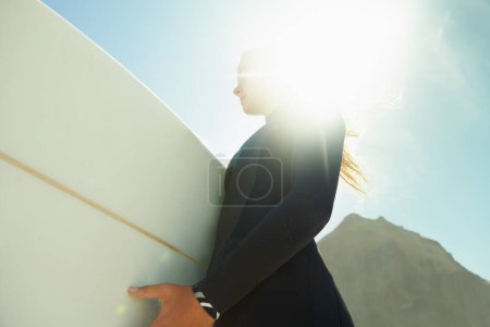 Foto de Bengala, cielo en la playa y mujer con tabla de surf en verano desde abajo para viajar, vacaciones o vacaciones. Ejercicio, fitness o deportes con un joven surfista confiado en traje de neopreno para el primer plano de ocio. - Imagen libre de derechos