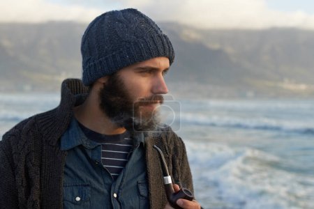 Photo for Beard, man and smoking a pipe by ocean, thinking and sailor habit on winter morning to relax. English person, nicotine and vintage smoker with breathe smoke, calm and vacation in cape town by beach. - Royalty Free Image
