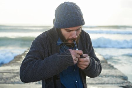 Photo for Bearded, man and smoking a pipe by ocean, lighter and tobacco habit on winter morning for sunrise. English guy, nicotine and vintage smoker for calm, satisfaction and vacation on beach in cape town. - Royalty Free Image