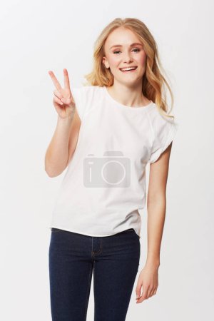 Foto de Mujer joven, signo de paz y retrato en estudio para la felicidad, positivo y funky en la moda casual. Modelo americano, sonrisa y cara con pelo rubio en jeans de moda, emoji y gen z por fondo blanco. - Imagen libre de derechos