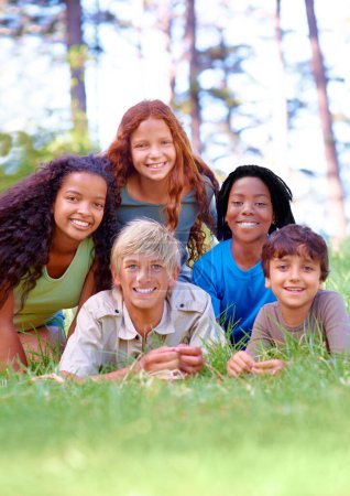 Téléchargez les photos : Nature, heureux et portrait d'enfants sur l'herbe dans le parc extérieur, champ ou jardin ensemble. Sourire, diversité et groupe de jeunes enfants excités se détendre et s'allonger sur la pelouse dans les bois ou la forêt pour l'été - en image libre de droit