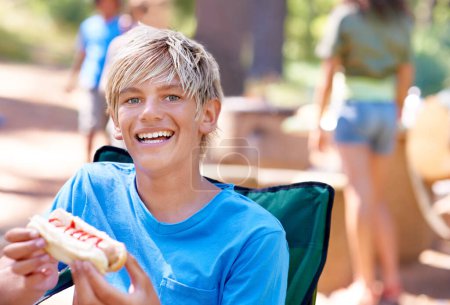 Téléchargez les photos : Manger, hot-dog et portrait d'enfant en plein air dans une chaise de camping et se détendre au barbecue avec le déjeuner. Heureux, enfant et avide de nourriture du barbecue dans le parc, les bois ou la forêt en vacances ou en vacances en été. - en image libre de droit