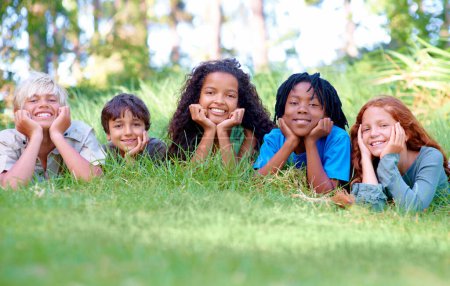Téléchargez les photos : Nature, sourire et portrait d'enfants sur l'herbe dans un parc extérieur, un champ ou un jardin ensemble. Heureux, diversité et groupe de jeunes enfants excités se détendre et s'allonger sur la pelouse dans les bois ou la forêt pour l'été - en image libre de droit