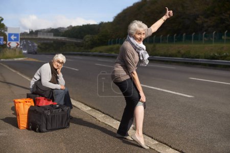 Photo for Women, road and funny hitch hiker for travel in retirement, lift and senior friends in city with bags for adventure. Elderly, thumbs up and tired by roadside with comic and stop transport to commute. - Royalty Free Image
