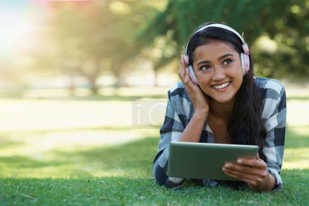 Photo for Woman, headphones and streaming podcast on tablet, learning and internet for education on grass in outdoors. Female person, happy and relaxing on lawn, knowledge and student subscription for info. - Royalty Free Image