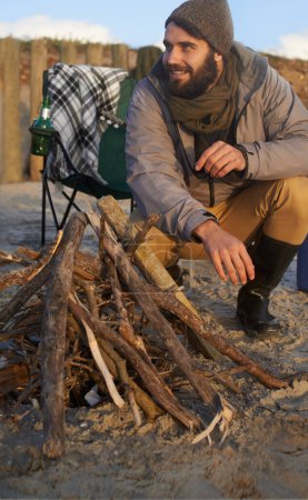 Photo for Campfire, wood and man by beach on vacation, adventure or holiday camping for travel. Nature, outdoor and young male person sitting on chair in sand with flame for heat on weekend trip in winter - Royalty Free Image