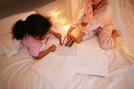 Téléchargez les photos : Les filles, crayon de couleur et dessin sur le lit pour la soirée pyjama, créatif et se détendre dans la chambre pour le week-end. Jeunes enfants, jeux ou jouets éducatifs la nuit en pyjama, amis ou collage par la lumière des fées à la maison. - en image libre de droit
