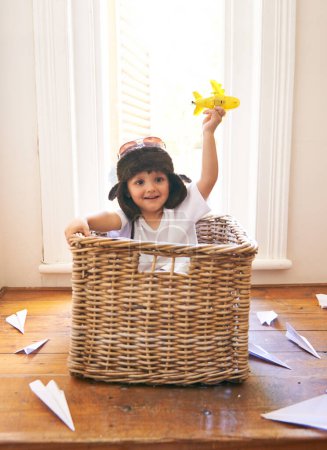 Photo for Child, smile and airplane toy in home, playful and freedom of youth with paper for fun and games. Little kid, happiness and hand in air with body in basket with hat on head as helmet with imagination. - Royalty Free Image