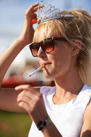 Téléchargez les photos : Couronne, fumeur et jeune femme avec cigarette dans la nature lors d'un festival, d'un événement ou d'une fête en plein air. Fun, tiare et femme avec du tabac, des lunettes de soleil et de la mode décontractée à l'extérieur du parc ou du champ - en image libre de droit