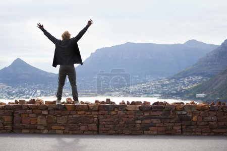 Photo for Person, watching and mountains with arms up in freedom and standing on brick wall in Cape Town. Excited, tourist or traveler with hands in the air for happiness on vacation, holiday or road trip. - Royalty Free Image