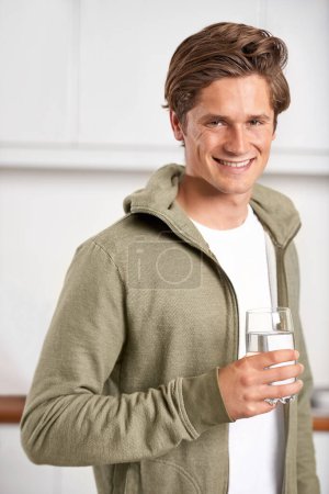 Foto de Sonrisa, retrato y hombre con vaso de agua en cocina de hogar para hidratación, salud y bienestar. Feliz, seguro y joven persona masculina beber líquido h2o fresco o bebida en apartamento moderno - Imagen libre de derechos