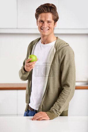 Photo for Smile, portrait and man with apple in kitchen of home for diet, health and wellness. Happy, confident and young male person eating fruit for organic, fresh and nutrition snack in modern apartment - Royalty Free Image