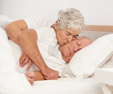 Photo for Senior couple, hug and kiss in bed or relaxing in retirement, love and bonding on weekend. Elderly people, sleeping man and embracing for affection in marriage, romance and morning routine at home. - Royalty Free Image