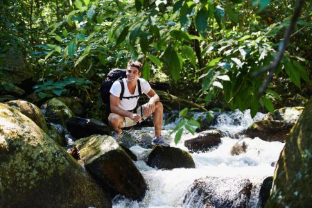 Téléchargez les photos : Homme, randonnée et rivière en forêt tropicale avec sourire pour l'aventure, le voyage et les rochers avec sac à dos. Personne, extérieur et sac par l'eau, la terre et les arbres dans la jungle en vacances avec liberté en Colombie. - en image libre de droit