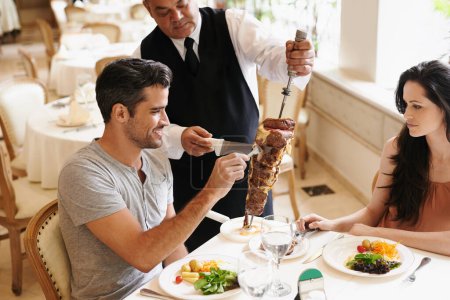 Photo for Happy couple, date and waiter with kebab for serving, fine dinning or romantic dinner at table. Young man and woman with chef cutting skewer of food for meal, eating or enjoying service at restaurant. - Royalty Free Image